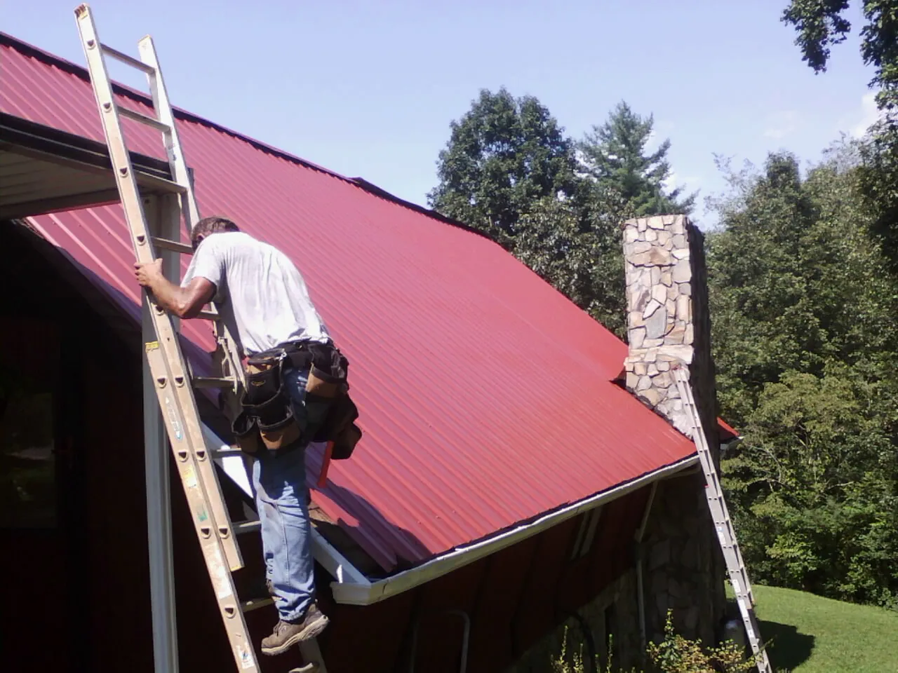 Roofer on a ladder beside a red metal roof and stone chimney — Soffit & Fascia Repair by Roofing Brothers in Onondaga